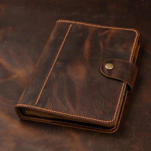 A dark brown leather planner or notebook cover with tan contrast stitching and a snap-button strap closure, photographed on a dark brown textured surface.
