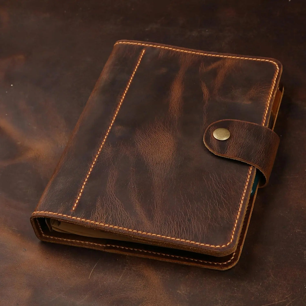 A dark brown leather planner or notebook cover with tan contrast stitching and a snap-button strap closure, photographed on a dark brown textured surface.
