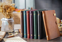 Set of leather-bound notebooks in various colors on a desk with a vase of dried flowers.