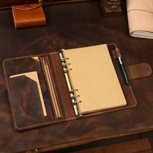 A brown leather ring binder notebook cover laid open on a dark brown wooden desk, holding a beige notebook with metal ring rings, card slots, a pen loop, and a small pouch. A matching brown leather accessory case is visible in the upper left, with other desk items partially shown around it.